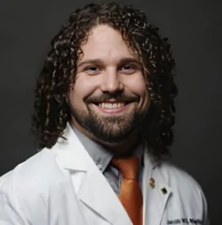 Professional headshot of male dentist in white coat and orange shirt against gray background.