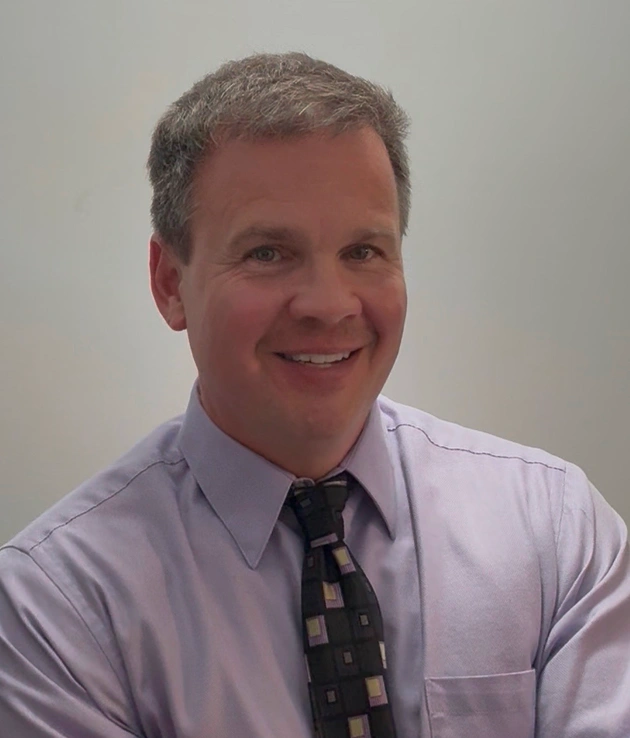 Professional headshot of a man in light purple shirt and tie smiling at camera in office setting.