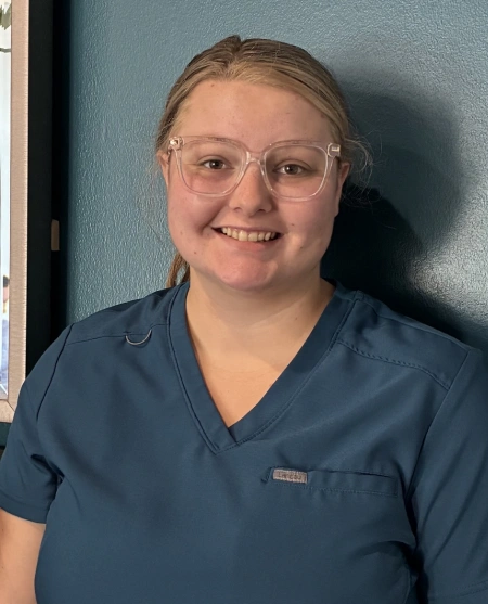 A medium close-up shot of a young woman with blonde hair tied back, wearing clear-framed glasses and a teal-colored V-neck medical scrub top. She is smiling directly at the camera, standing against a dark teal-blue wall with a framed picture visible on the left edge.