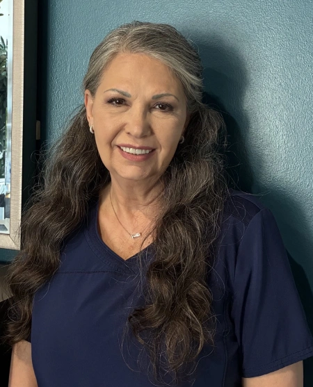 A medium close-up shot of a middle-aged woman with long, wavy, dark hair streaked with grey, wearing a dark navy blue V-neck medical scrub top and a delicate silver necklace. She is smiling at the camera, standing against a dark teal-blue wall with a framed picture visible on the left edge.