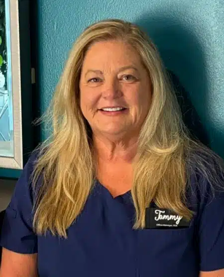 Professional dental staff member smiling at camera wearing navy scrubs with name badge in clinical setting.